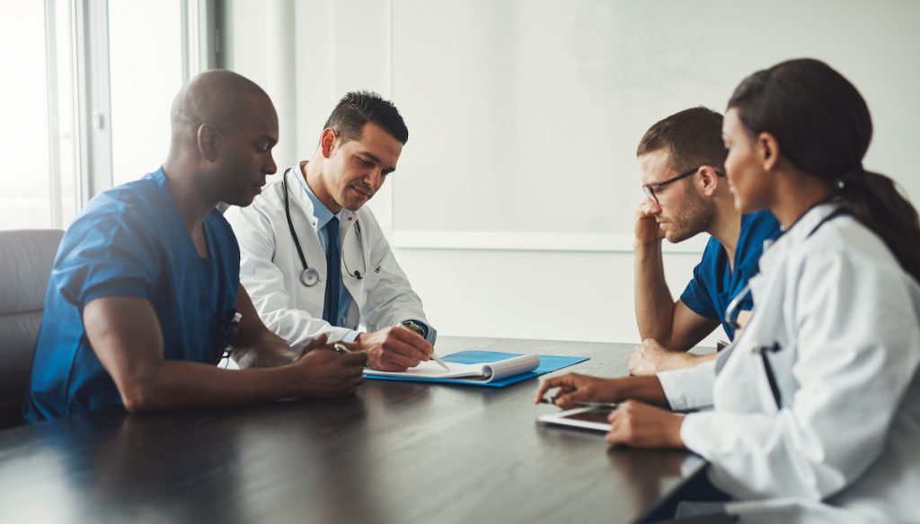 Multiracial medical team having a meeting with doctors in white lab coats and surgical scrubs seated at a table discussing a patients records