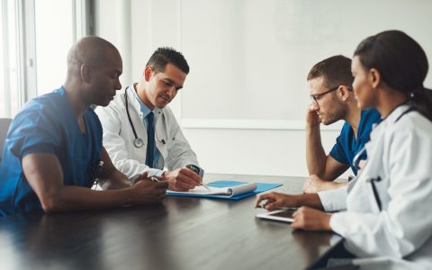 Multiracial medical team having a meeting with doctors in white lab coats and surgical scrubs seated at a table discussing a patients records