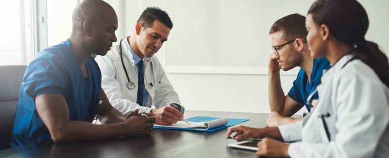 Multiracial medical team having a meeting with doctors in white lab coats and surgical scrubs seated at a table discussing a patients records